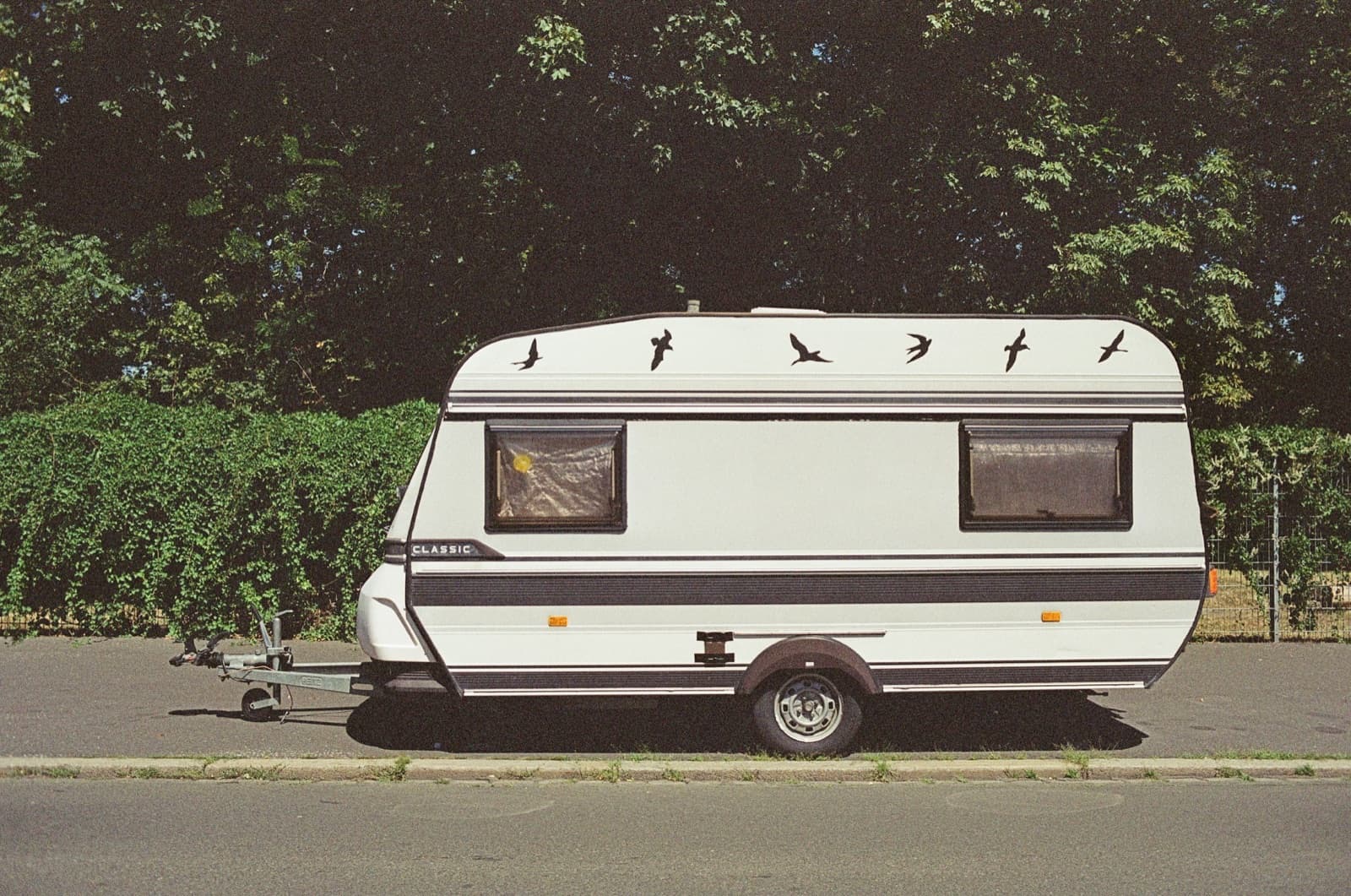 Une caravane ancienne à un essieu stationnée au bord d'une rue, devant une haie.